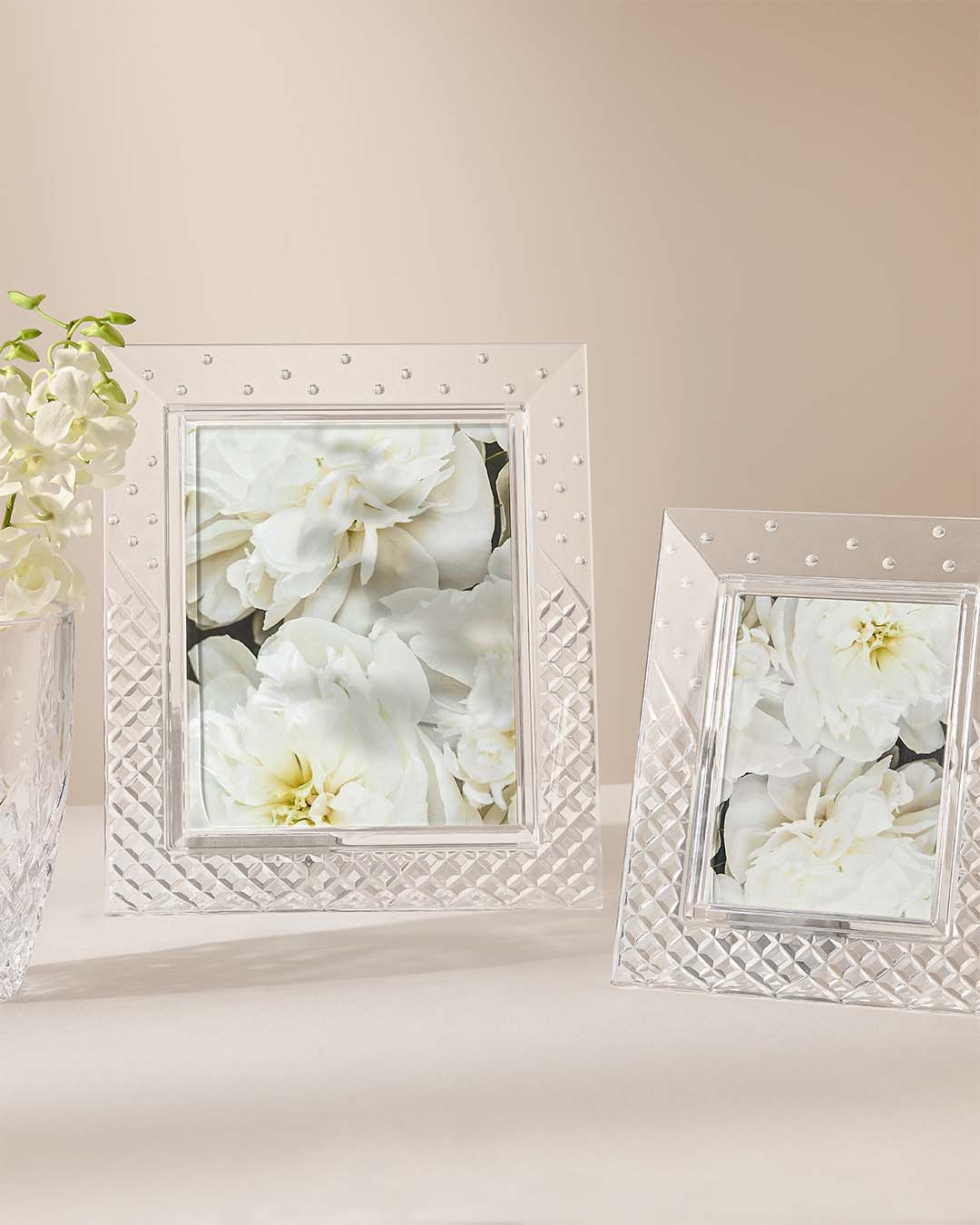Two clear textured picture frames with small bead accents, one standing upright and one leaning, both displaying a white flower photo. A vase of white flowers sits on the left against a soft beige background.