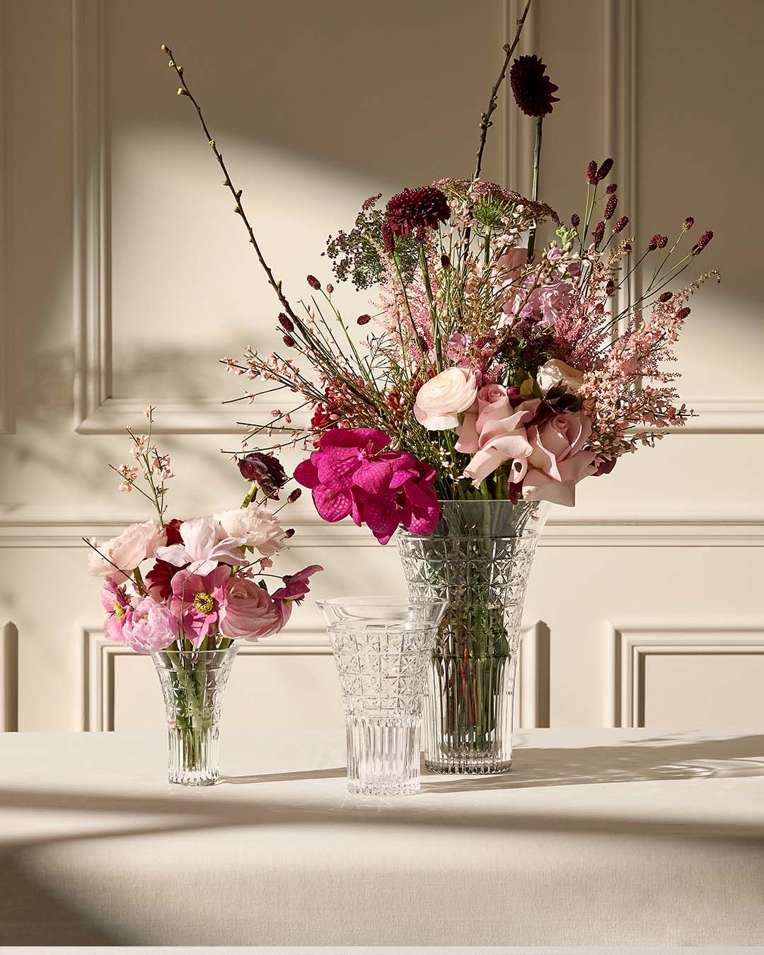 Three clear cut-glass flower vases arranged on a cream table against a paneled wall, holding pink, burgundy, and mauve floral arrangements with sunlight and shadows.