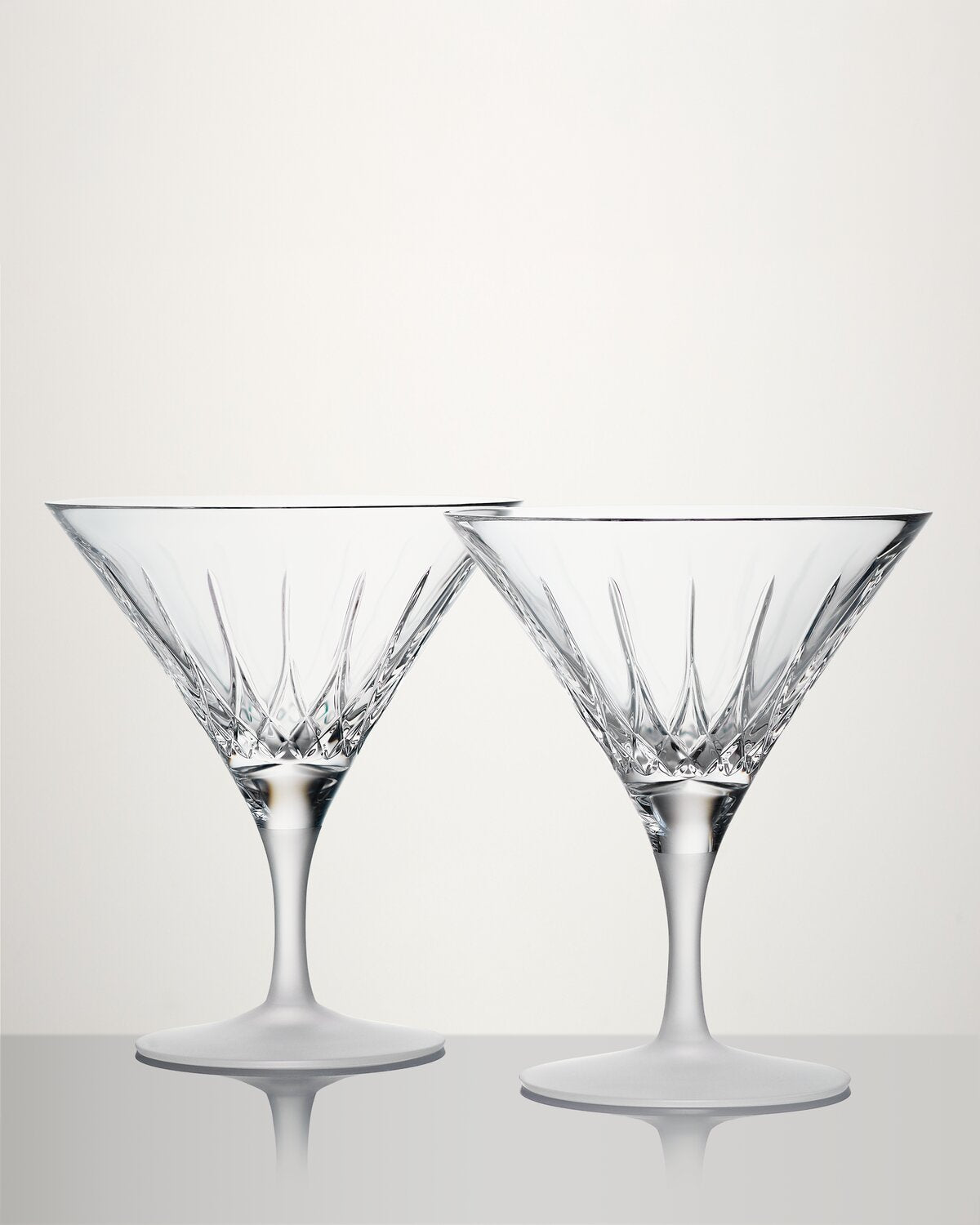 Two clear cut-glass martini glasses with faceted stems and circular bases, photographed on a light gray studio background with a reflective surface below.