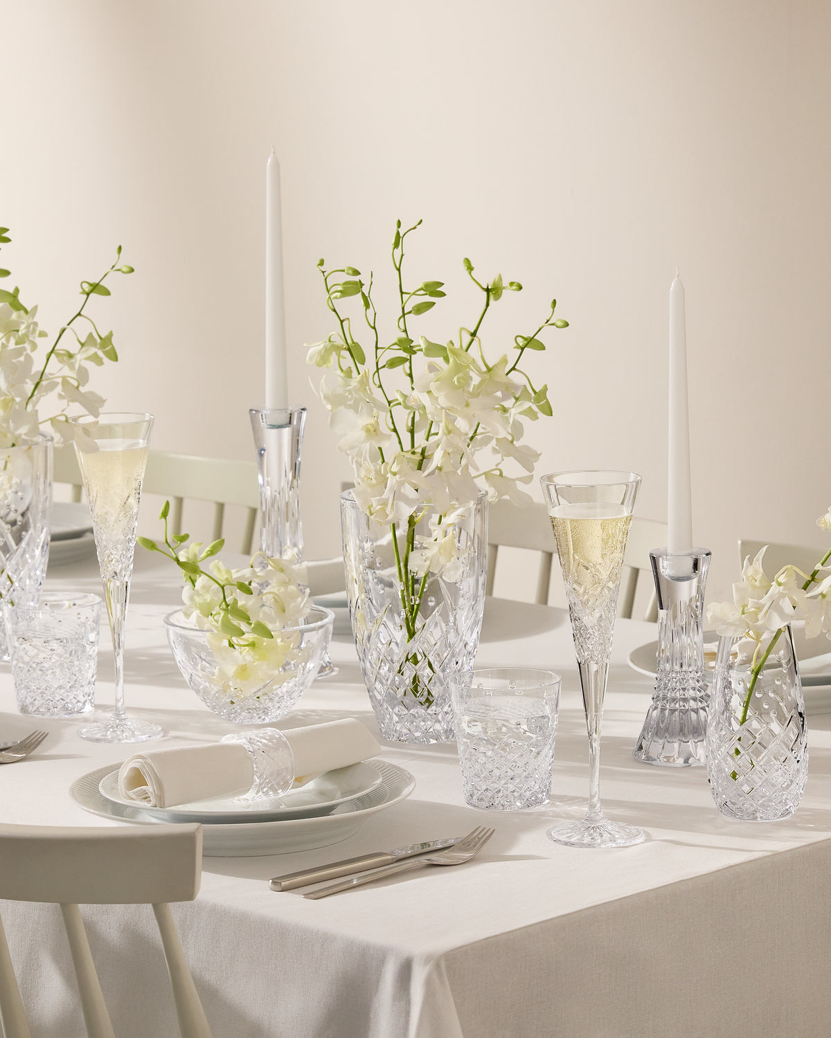 A beautifully set dining table with clear crystal glassware, white plates, folded napkins, silver cutlery, and white orchid flower arrangements with tall white taper candles against a soft cream background.