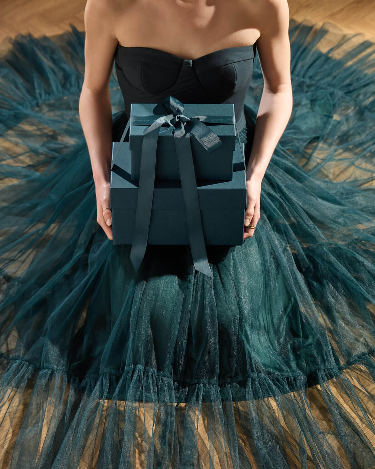 A model wearing a strapless black corset top and a voluminous dark teal tulle skirt holds a stack of dark teal gift boxes tied with a ribbon.