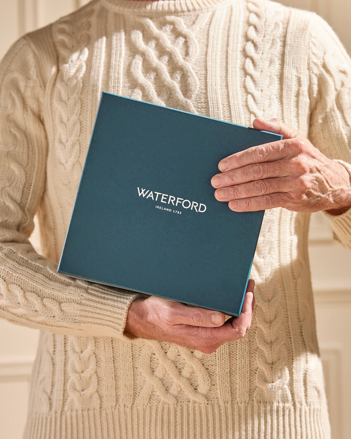 A person in a cream cable-knit sweater holds a dark teal Waterford gift box with white lettering, set against a softly lit beige background.