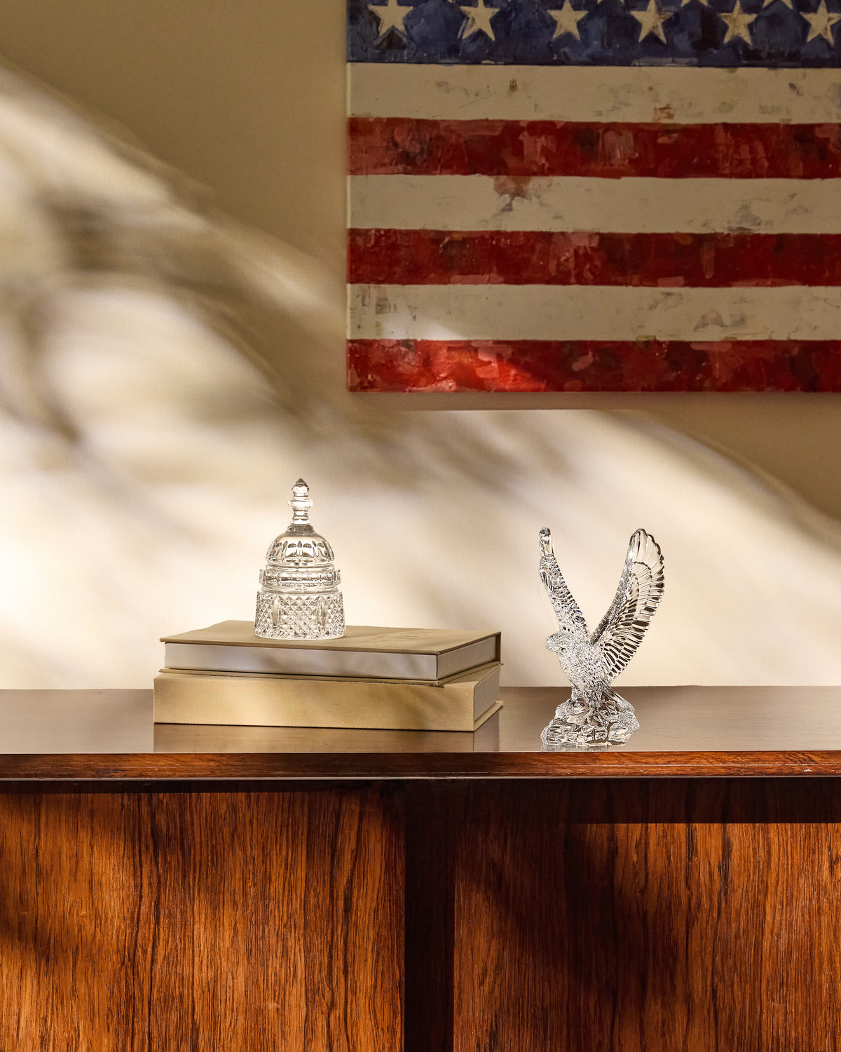 A silver-toned crystal eagle figurine and a small crystal dome-shaped ornament displayed on stacked books atop a wooden console table, with a distressed American flag hanging on the wall behind.